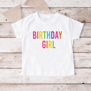 May include: White t-shirt with the text "BIRTHDAY GIRL" in rainbow colors.