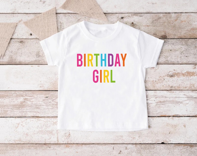May include: White t-shirt with the text "BIRTHDAY GIRL" in rainbow colors.