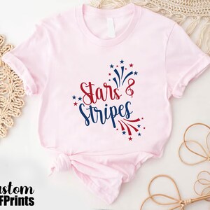 May include: A pink t-shirt with a red, white, and blue graphic that says "Stars & Stripes". The graphic features a firework design.