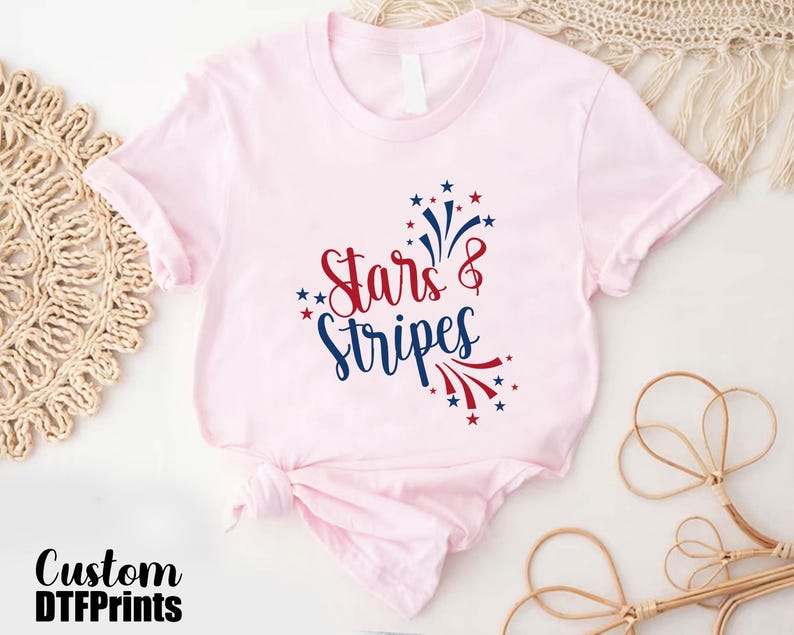 May include: A pink t-shirt with a red, white, and blue graphic that says "Stars & Stripes". The graphic features a firework design.