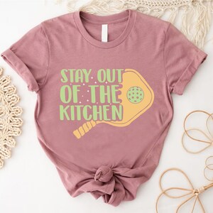 May include: A mauve t-shirt with a graphic of a paddleball and the text "Stay Out of the Kitchen".