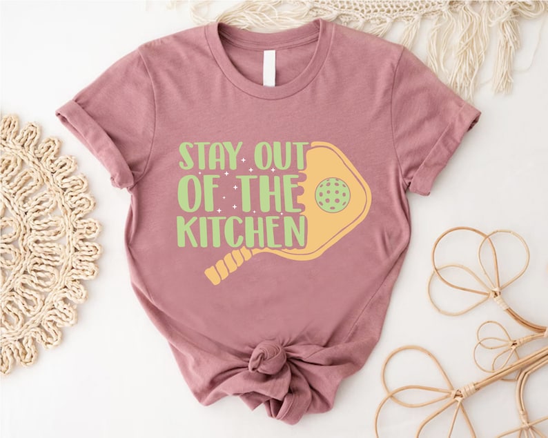 May include: A mauve t-shirt with a graphic of a paddleball and the text "Stay Out of the Kitchen".