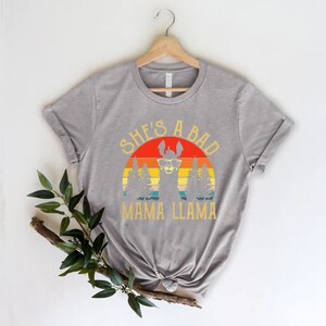 May include: Gray t-shirt with a retro sunset graphic featuring a llama wearing sunglasses. The text "She's a Bad Mama Llama" is printed below the graphic.