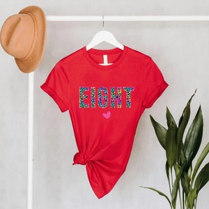 May include: Red short-sleeved t-shirt with the word "EIGHT" printed in a colourful leopard print design. A small pink heart is below the word.