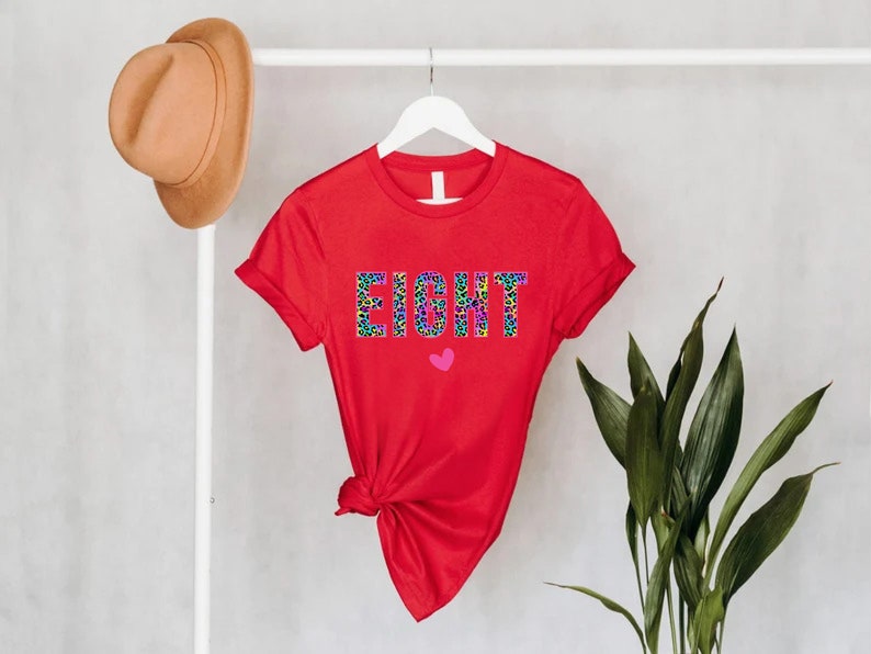 May include: Red short-sleeved t-shirt with the word "EIGHT" printed in a colourful leopard print design. A small pink heart is below the word.