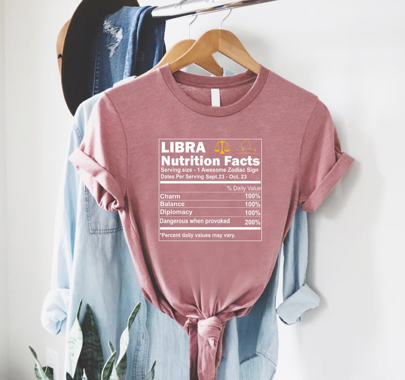 May include: A pink t-shirt with a graphic of a nutrition facts label for the zodiac sign Libra. The label lists the daily value percentages for charm, balance, diplomacy, and dangerous when provoked. The text "Libra Nutrition Facts" is at the top of the label.