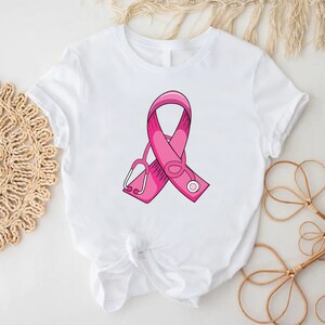 May include: White t-shirt with a pink ribbon and a stethoscope design. The ribbon is a symbol of breast cancer awareness.