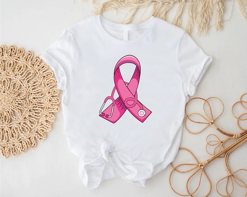 May include: White t-shirt with a pink ribbon and a stethoscope design. The ribbon is a symbol of breast cancer awareness.