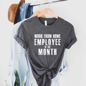 May include: Dark gray t-shirt with the text "WORK FROM HOME EMPLOYEE OF THE MONTH" in white block letters. The shirt is hanging on a wooden hanger with other clothing items in the background.