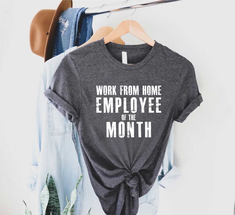 May include: Dark gray t-shirt with the text "WORK FROM HOME EMPLOYEE OF THE MONTH" in white block letters. The shirt is hanging on a wooden hanger with other clothing items in the background.