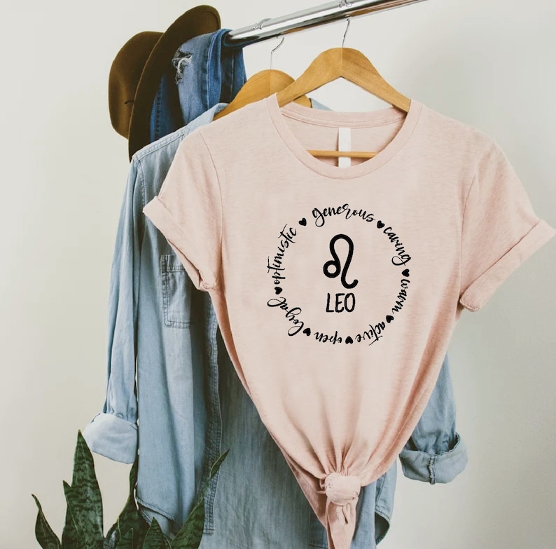 May include: A pink t-shirt with a black circle design featuring the Leo zodiac symbol and the words "Leo", "optimistic", "generous", "caring", "loyal", "active", "open", "brave".
