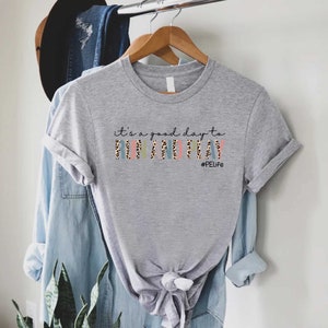 K&ouml;nnte beinhalten: Graues T-Shirt mit dem Text "it's a good day to run and play" in Schwarz mit Leopardenmuster. Der Text "#PELife" ist in kleinerer Schrift unter dem Haupttext.