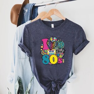 May include: A dark blue t-shirt featuring a graphic print of a heart with a leopard print design and the text "I love the 80's" surrounded by 80s themed icons.