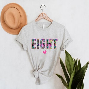 May include: A grey t-shirt with the word "EIGHT" printed in rainbow leopard print with a pink heart below.