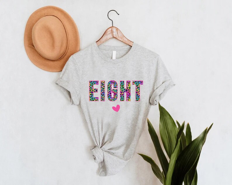 May include: A grey t-shirt with the word "EIGHT" printed in rainbow leopard print with a pink heart below.