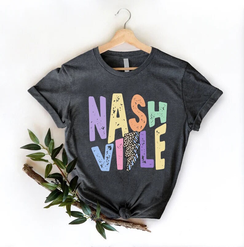 Puede incluir: Una camiseta gris oscuro con un gr&aacute;fico colorido que dice "NASHVILLE" con un rayo en el medio de la "I".
