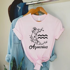May include: A pink t-shirt with a black graphic of a crescent moon with a floral design and the word "Aquarius" underneath.