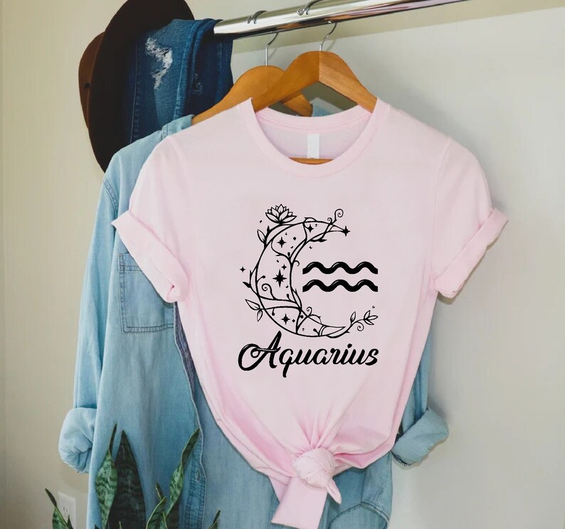 May include: A pink t-shirt with a black graphic of a crescent moon with a floral design and the word "Aquarius" underneath.