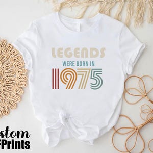 May include: White t-shirt with a vintage-style graphic that says "Legends were born in 1975". The graphic is in shades of brown, orange, and yellow.
