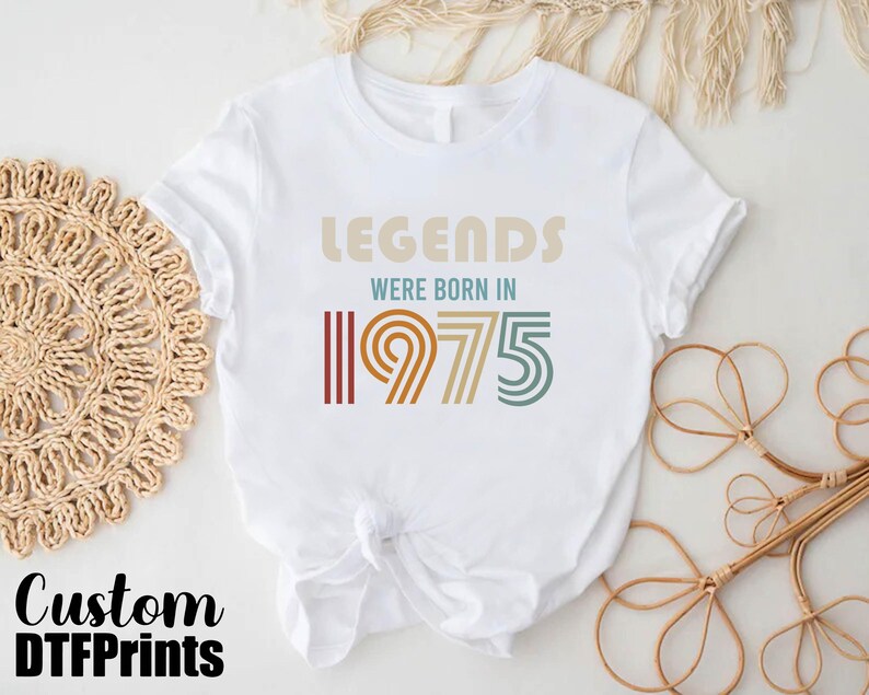 May include: White t-shirt with a vintage-style graphic that says "Legends were born in 1975". The graphic is in shades of brown, orange, and yellow.