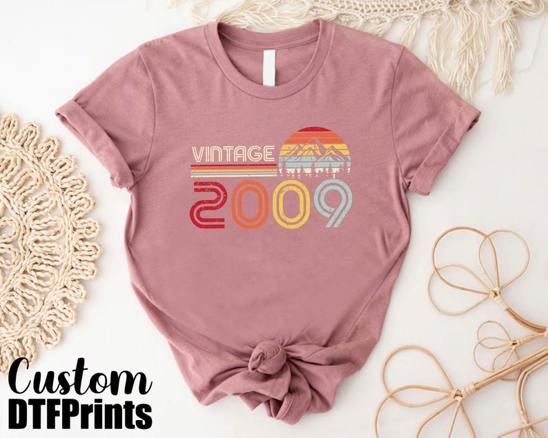 May include: A pink t-shirt with a vintage design featuring a mountain range and the text "Vintage 2009".