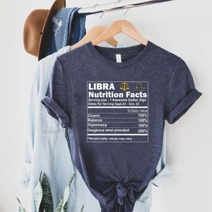 May include: A dark blue t-shirt with a white graphic of a nutrition facts label for the zodiac sign Libra. The label includes the text "LIBRA Nutrition Facts" and lists the following: Charm, Balance, Diplomacy, Dangerous when provoked. Each item has a percentage daily value listed next to it.