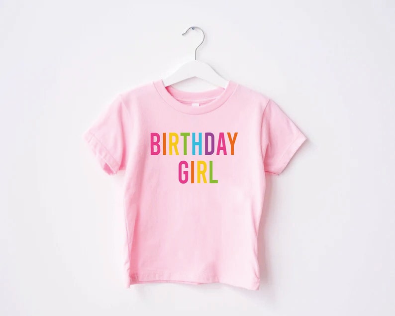 May include: Pink short-sleeved t-shirt with the text "BIRTHDAY GIRL" in rainbow colors.