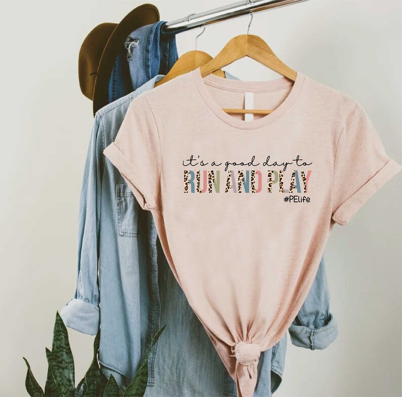 K&ouml;nnte beinhalten: Ein pinkfarbenes T-Shirt mit dem Text "it's a good day to RUN AND PLAY #PELife" in bunten Buchstaben mit Leopardenmuster.