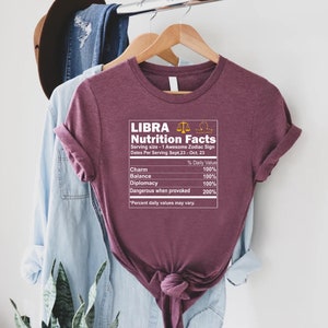 May include: A maroon t-shirt with a graphic of a nutrition facts label for the zodiac sign Libra. The label lists the daily value of charm, balance, diplomacy, and dangerous when provoked. The text reads "Libra Nutrition Facts Serving size - 1 Awesome Zodiac Sign Dates Per Serving Sept.23 - Oct. 23 % Daily Value Charm 100% Balance 100% Diplomacy 100% Dangerous when provoked 200% "Percent daily values may vary."