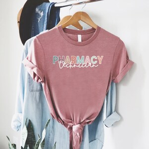 May include: A pink t-shirt with the text "PHARMACY Technician" in colorful letters. The shirt is hanging on a wooden hanger.