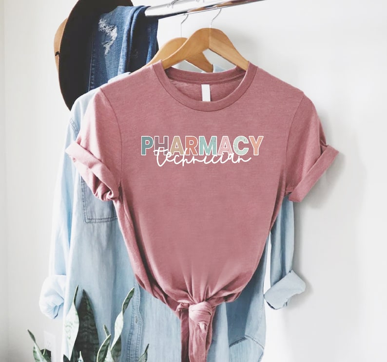 May include: A pink t-shirt with the text "PHARMACY Technician" in colorful letters. The shirt is hanging on a wooden hanger.