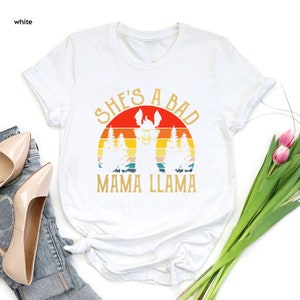 May include: White t-shirt with a retro sunset graphic and a llama wearing sunglasses. The text "She's a Bad Mama Llama" is printed on the shirt.