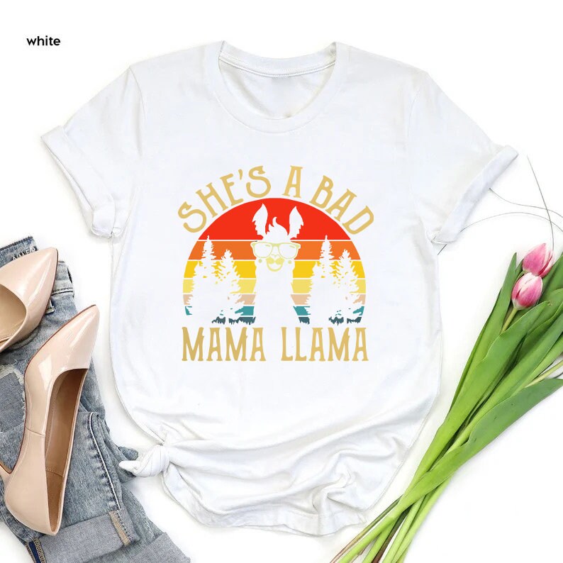 May include: White t-shirt with a retro sunset graphic and a llama wearing sunglasses. The text "She's a Bad Mama Llama" is printed on the shirt.