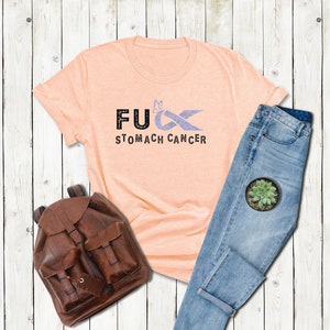 May include: A peach-colored t-shirt with the text "F**K STOMACH CANCER" and a purple ribbon with a butterfly on it.