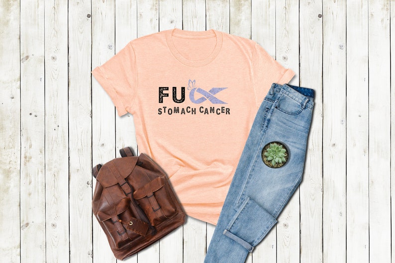 May include: A peach-colored t-shirt with the text "F**K STOMACH CANCER" and a purple ribbon with a butterfly on it.