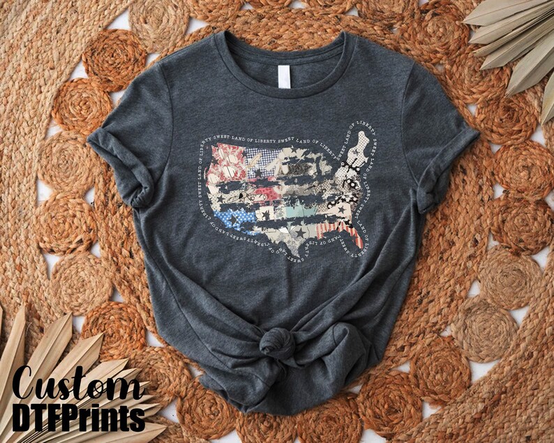 May include: A dark gray t-shirt with a distressed American flag design featuring a patchwork of red, white, and blue fabric. The design is surrounded by the text "Sweet Land of Liberty Sweet Land of Liberty Sweet Land of Liberty Sweet Land of Liberty".