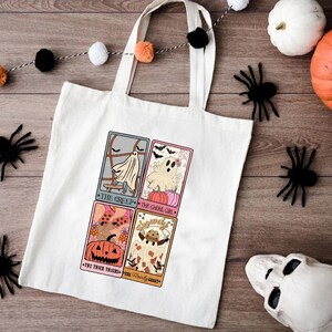 May include: A white tote bag featuring four illustrated cards with Halloween themes. The cards depict a ghost, a pumpkin, a witch, and a friendly ghost. The text on the cards reads "The Creep", "The Ghoul Girl", "The Trick Treats", and "The Friendly Ghost".