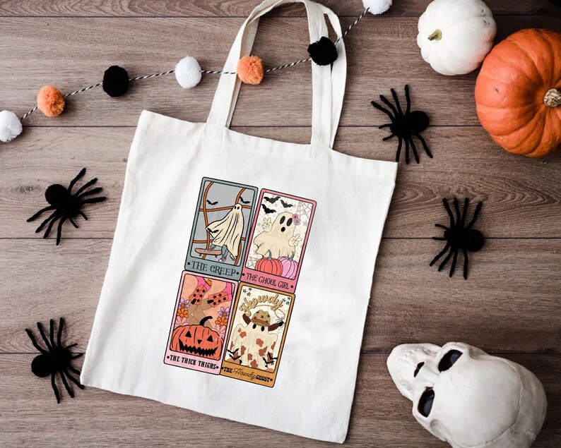 May include: A white tote bag featuring four illustrated cards with Halloween themes. The cards depict a ghost, a pumpkin, a witch, and a friendly ghost. The text on the cards reads "The Creep", "The Ghoul Girl", "The Trick Treats", and "The Friendly Ghost".