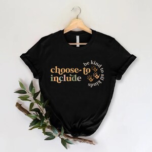 以下が含まれることがあります： オレンジ、緑、ベージュで「choose to include」という言葉が書かれた黒いTシャツ。また、このシャツには「be kind to all kinds」というフレーズと手のひらのプリントが円で描かれています。