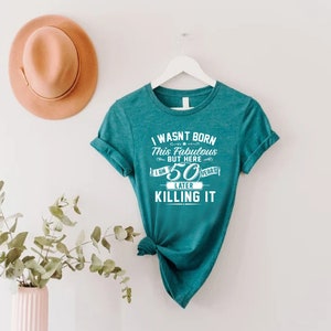 Pode incluir: Camiseta azul turquesa com texto branco que diz "Eu n&atilde;o nasci assim, mas aqui estou, 50 anos depois e estou arrasando".