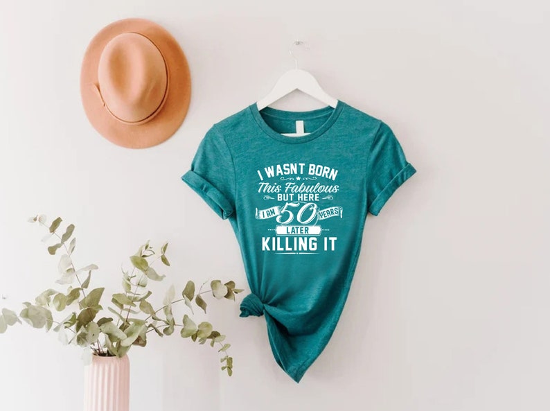 Pode incluir: Camiseta azul turquesa com texto branco que diz "Eu n&atilde;o nasci assim, mas aqui estou, 50 anos depois e estou arrasando".