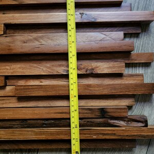 May include: A close-up of a wooden wall panel made of various sized rectangular pieces of wood. The panel is approximately 12 inches wide and 13 inches tall. A yellow measuring tape is placed next to the panel.
