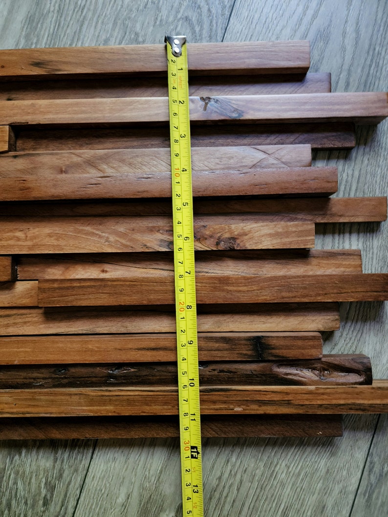 May include: A close-up of a wooden wall panel made of various sized rectangular pieces of wood. The panel is approximately 12 inches wide and 13 inches tall. A yellow measuring tape is placed next to the panel.