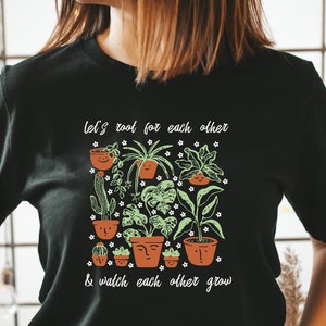 May include: Black t-shirt with a white graphic of potted plants with faces. The text on the shirt reads "Let's root for each other & watch each other grow."