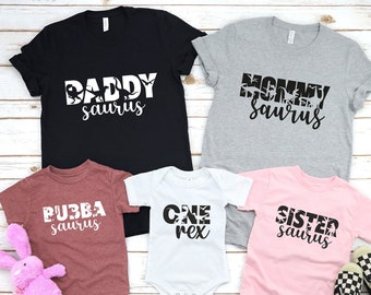 May include: A collection of family dinosaur-themed t-shirts and a onesie. The shirts are black, gray, maroon, and pink, with the words "Daddy Saurus", "Mommy Saurus", "Bubba Saurus", and "Sister Saurus". The white onesie says "One Rex".