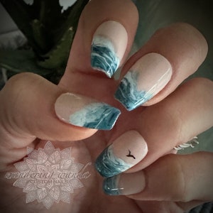 May include: Close-up of a hand with blue and white ocean wave nail art. The nails are painted with a light pink base and have a blue and white marbled design that resembles ocean waves. One nail features a small black silhouette of a bird flying over the waves.