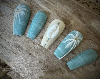 May include: A set of custom-designed, long, coffin-shaped false nails. The nails feature a variety of designs, including a seashell, palm trees, and textured patterns in shades of blue, white, and beige. The nails are arranged on a wooden surface.