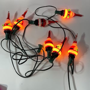 Op de afbeelding: Een kerstverlichtingssnoer met rode, witte en groene kerstman-lampjes. De lampjes zijn aan en gloeien oranje.