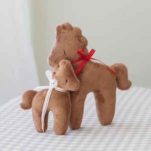 Può includere: Due giocattoli di peluche a forma di cavallo marrone, uno più grande e uno più piccolo, affiancati. Il cavallo più grande ha un nastro rosso legato al collo, mentre il cavallo più piccolo ha un nastro bianco. Sono su una superficie a scacchi.
