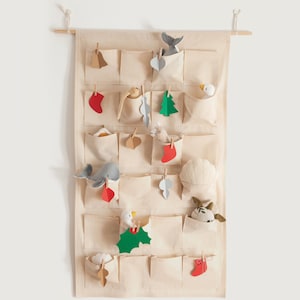 Pode incluir: Um calendário do advento de tecido bege com 24 bolsos, cada um com um pequeno brinquedo ou decoração. Os bolsos estão cheios de uma variedade de itens, incluindo uma baleia, um pássaro, uma estrela, uma árvore e uma meia.